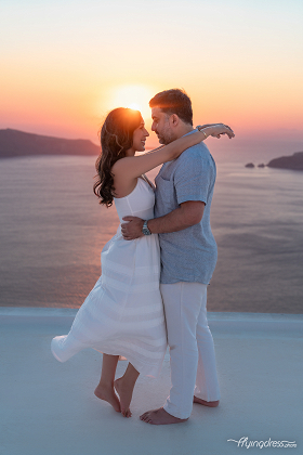 A couple embraces on a rooftop in Santorini during a sunset, with the sun setting directly behind them, creating a soft romantic glow. The woman's light dress and the serene sea in the background enhance the atmosphere of love and tranquility.