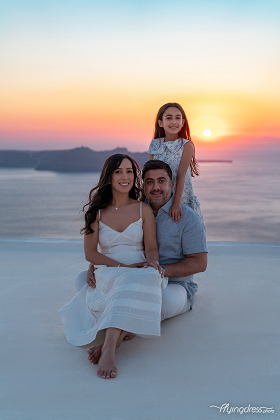 A family poses at sunset in Santorini, seated on a rooftop with the ocean and cliffs in the background. The warm hues of the sky enhance the serene, joyful moment.