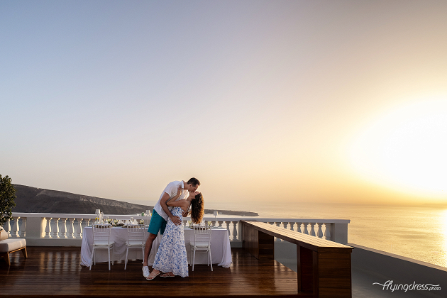 A couple shares a romantic kiss at sunset on a beautifully set terrace overlooking the ocean, creating a dreamy and intimate scene.