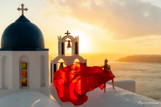 A woman in a flowing red dress poses at sunset beside a whitewashed church with blue domes in Santorini, Greece.