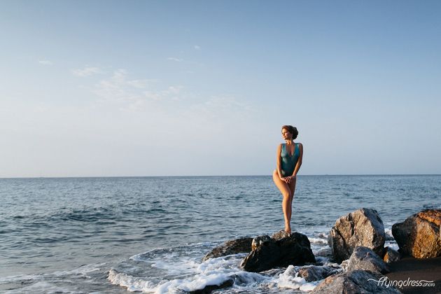 Standing tall on the rocky black beach of Santorini, a girl in a swimsuit embraces the untamed allure of the volcanic shores, surrounded by the dramatic contrast of the dark sands and the endless expanse of the azure sea.