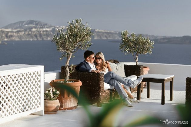 A couple shares a cozy moment, seated on a stylish sofa, their connection evident through their warm smiles and intertwined hands, as they create a snapshot of love and comfort in the picturesque setting of Santorini.