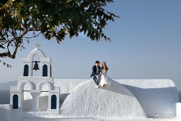 A couple sits intimately on a picturesque church in Santorini, their embrace blending the serenity of the sacred space with the depth of their love, creating a timeless moment of connection and devotion.