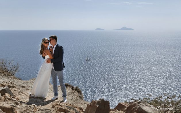A wedding couple stands hand in hand at the iconic lighthouse of Santorini, surrounded by the sweeping vistas of the Aegean Sea, as their love shines brightly amidst the timeless beauty of this romantic setting.
