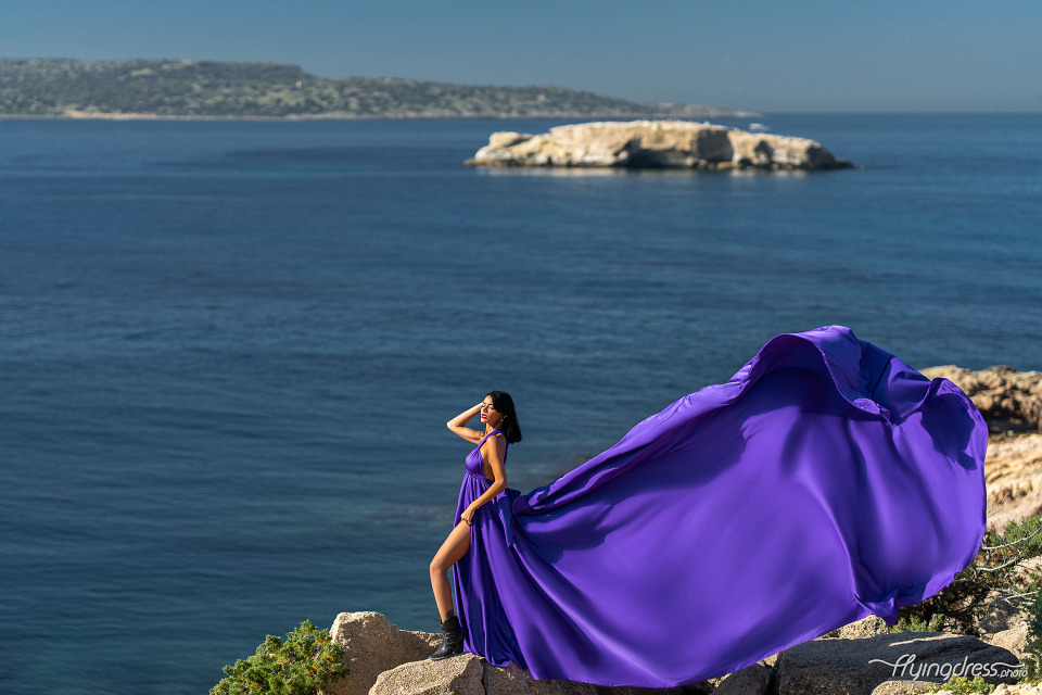 A woman in a flowing purple dress and black boots stands elegantly on rocky cliffs with the vast blue sea and distant islands of Kavouri, Athens in the background, creating a striking and graceful scene.