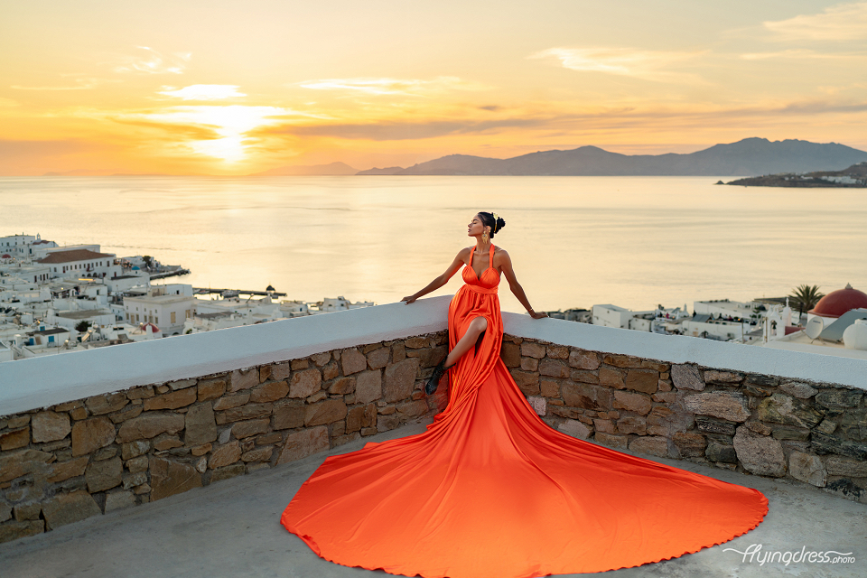 A woman in a flowing, bright orange dress poses against a stone wall with a stunning sunset and the town of Mykonos in the background.