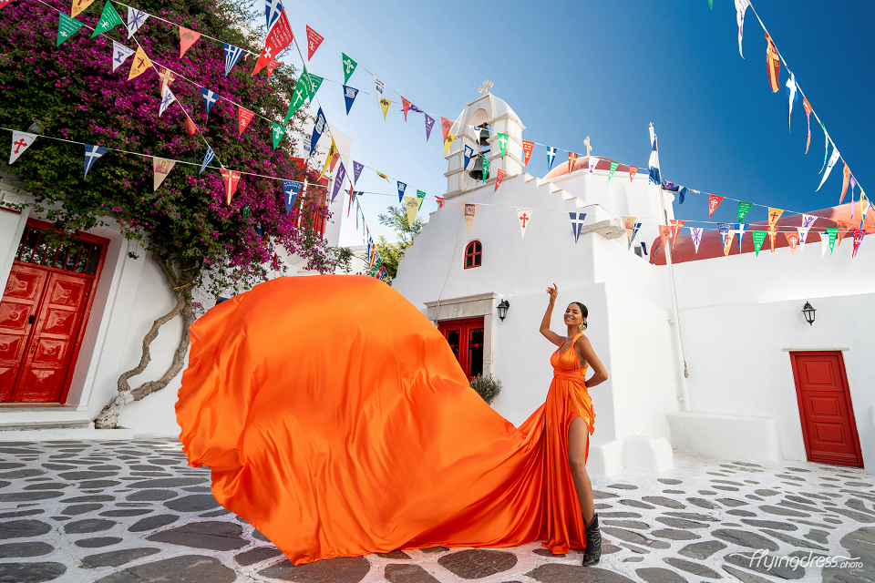 Celebrate the vibrant charm of Mykonos Chora with a flying dress photoshoot, where the striking orange gown beautifully complements the festive flags and white-washed architecture.