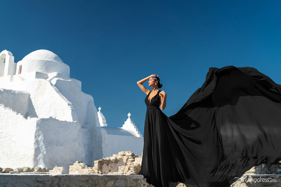 A woman in a flowing black dress poses against the backdrop of the iconic Paraportiani Church in Mykonos, with its whitewashed walls and a clear blue sky.