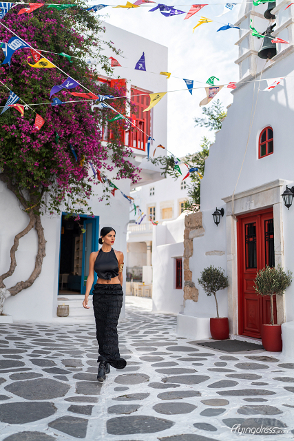 A woman in a chic black outfit walks confidently through a picturesque alley in Mykonos, surrounded by white-washed buildings, vibrant red doors, blooming bougainvillea, and colorful flags fluttering in the breeze.