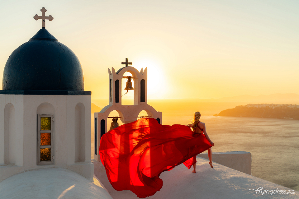 A woman in a flowing red dress poses at sunset beside a whitewashed church with blue domes in Santorini, Greece.