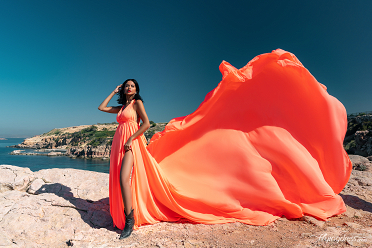 A woman in a vibrant orange flowing dress and black boots stands confidently on rocky cliffs with the scenic coastline and deep blue waters of Kavouri, Athens in the background.