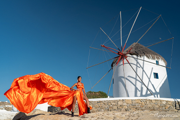 A woman in a vibrant orange gown poses dramatically in front of the iconic windmills of Mykonos, with the flowing dress contrasting beautifully against the clear blue sky and whitewashed structures.