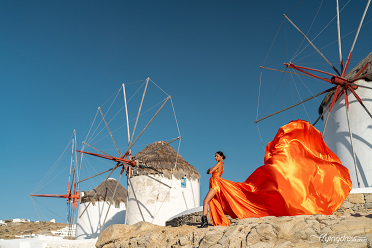 A woman in a flowing orange gown poses confidently in front of the iconic windmills of Mykonos, with her dress billowing dramatically against the clear blue sky and the historic structures providing a stunning backdrop.