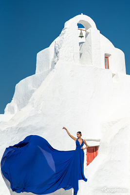 A woman in a flowing blue dress poses gracefully in front of the Paraportiani Church in Mykonos, with the wind catching her dress and the white structure contrasting against the clear blue sky.