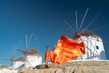 A woman in a flowing orange dress poses in front of a traditional windmill on Mykonos, with a clear blue sky and the wind catching her dress dramatically.