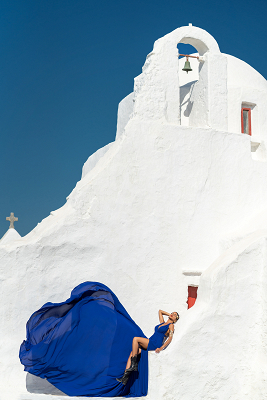 Experience the captivating allure of Mykonos with a flying dress photoshoot at the Church of Panagia Paraportiani, where the striking blue gown contrasts beautifully against the iconic white architecture and deep blue sky.