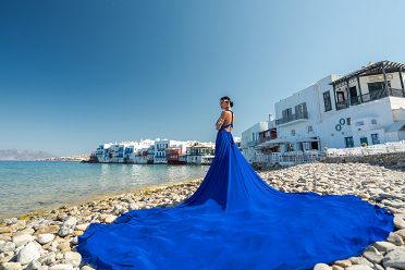 A woman in a flowing blue gown poses elegantly on the rocky shoreline of Mykonos, with the vibrant waterfront buildings of Little Venice and the clear Aegean Sea as a stunning backdrop.