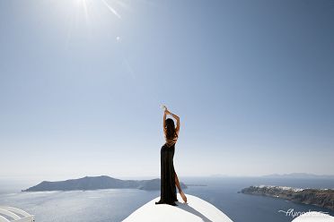 Standing against the backdrop of Santorini's breathtaking caldera, a girl in an elegant black dress radiates confidence and allure, perfectly complementing the island's captivating beauty and evoking a sense of timeless charm amidst the enchanting Mediterranean ambiance.