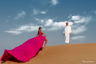 In the heart of Dubai's desert, a couple shares an intimate moment, the wind gently lifting a vibrant fuchsia flying dress as they capture the essence of love against the golden sands and endless horizons.