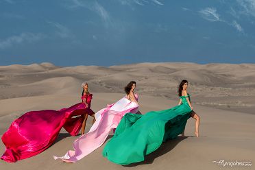 Three models radiate sophistication in a flying dress photoshoot amidst the timeless beauty of Dubai's desert, their dresses flowing gracefully in the wind against the backdrop of golden sands and endless horizons.